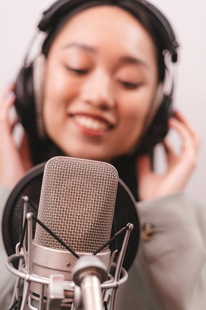 An Asian woman in a recording studio singing into a condenser microphone, wearing headphones.